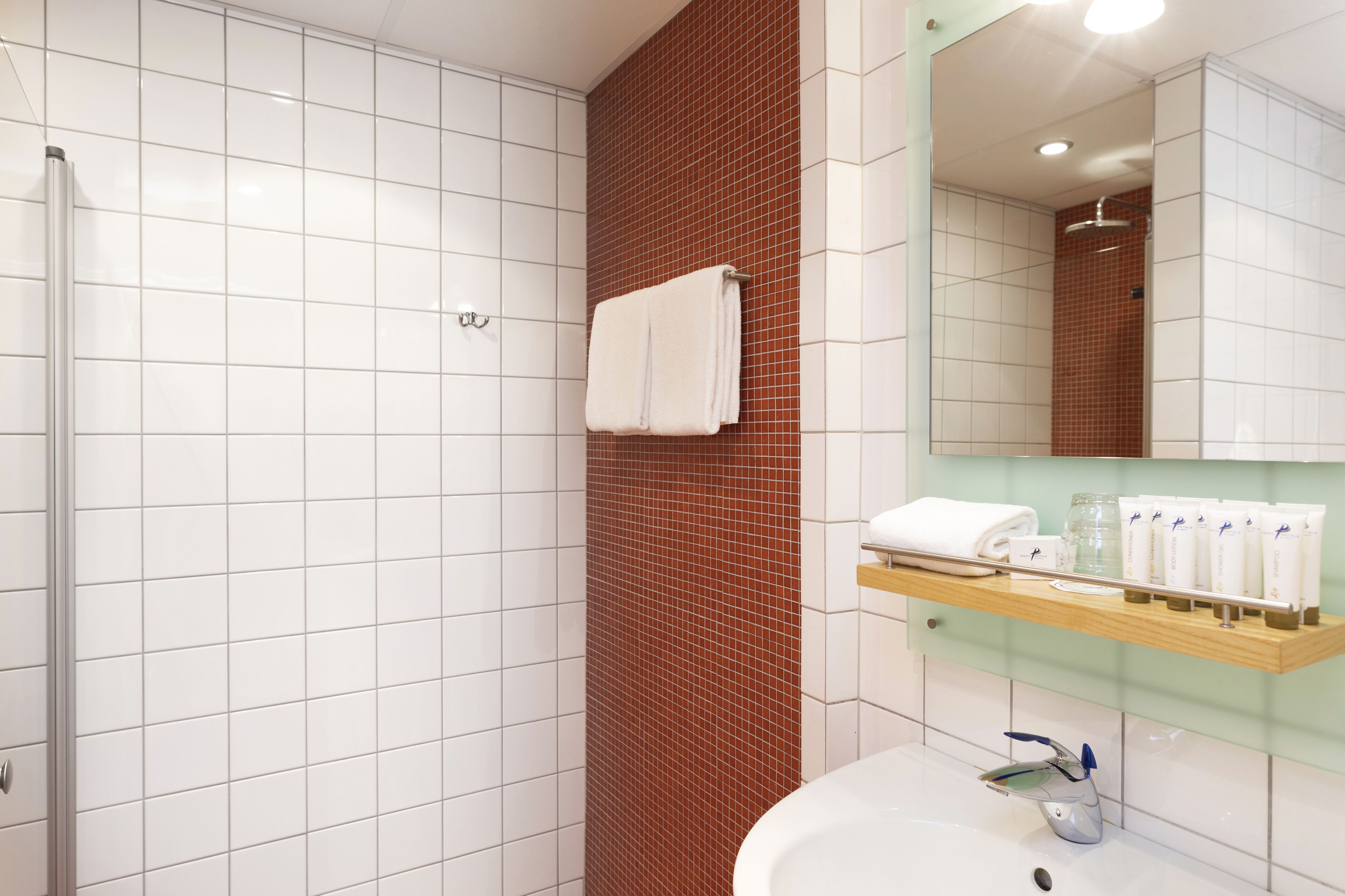 A bathroom with white tiles, a red-tiled shower wall, a glass shelf with toiletries, and a mirror.