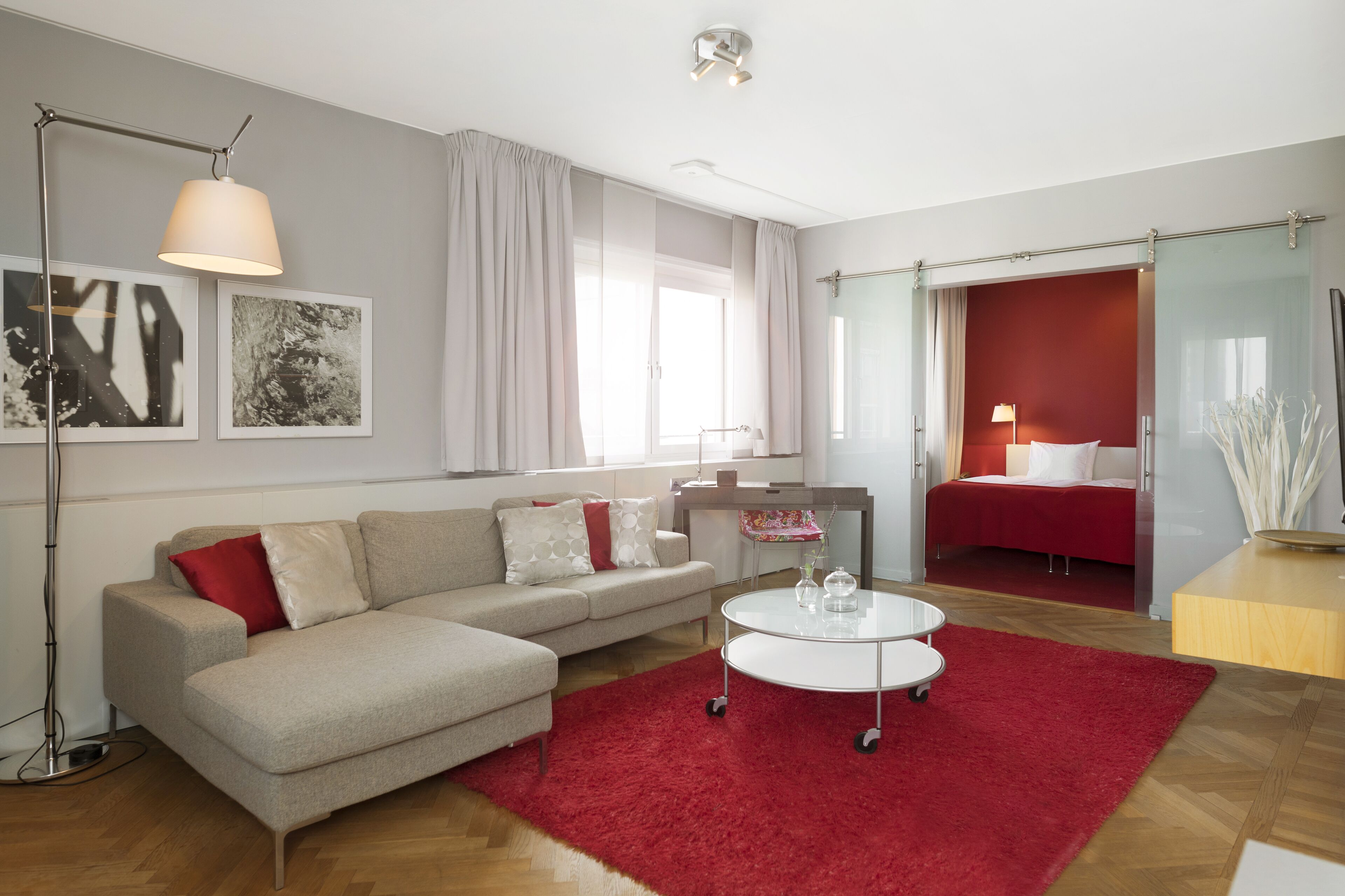 Una sala de estar moderna con un sofá, una mesa de centro y una alfombra roja. Se ve una cama al fondo y una ventana con cortinas.