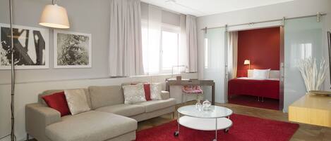 Una sala de estar moderna con un sofá, una mesa de centro y una alfombra roja. Se ve una cama al fondo y una ventana con cortinas.