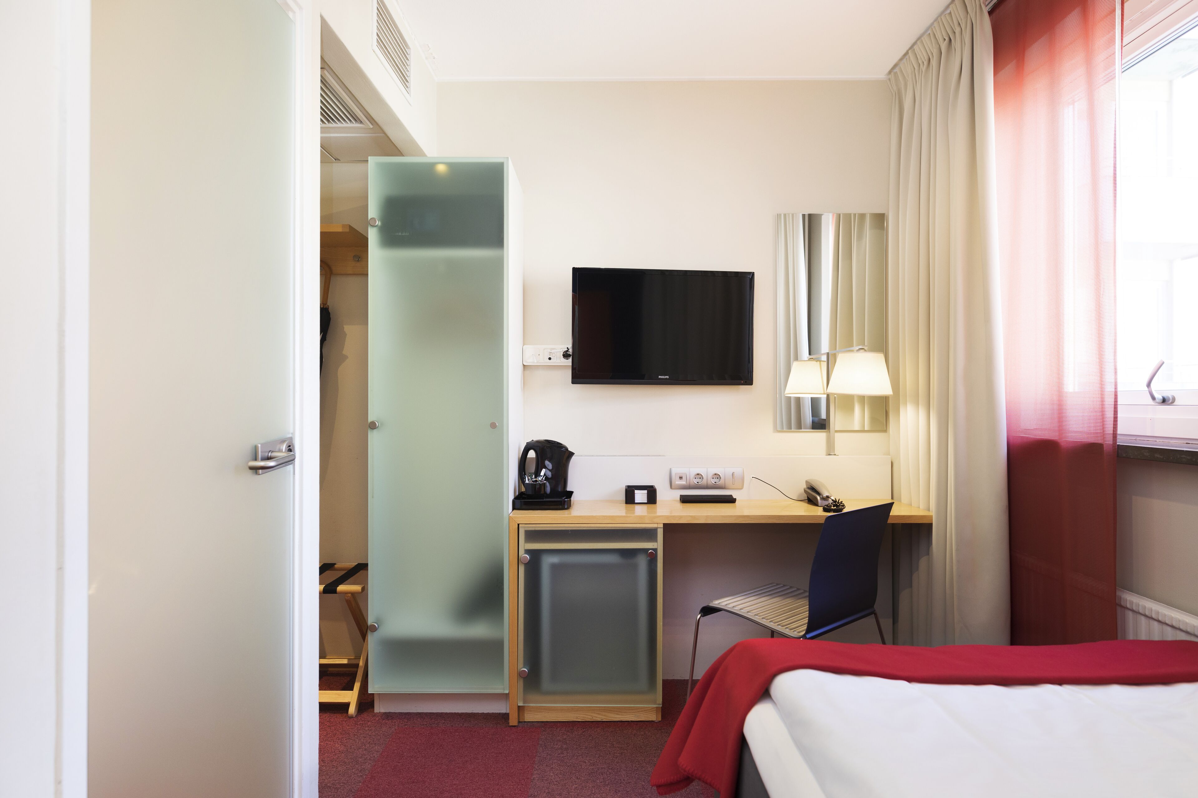 Habitación de hotel con cama, escritorio con silla, televisión, refrigerador pequeño y ventana con cortinas.