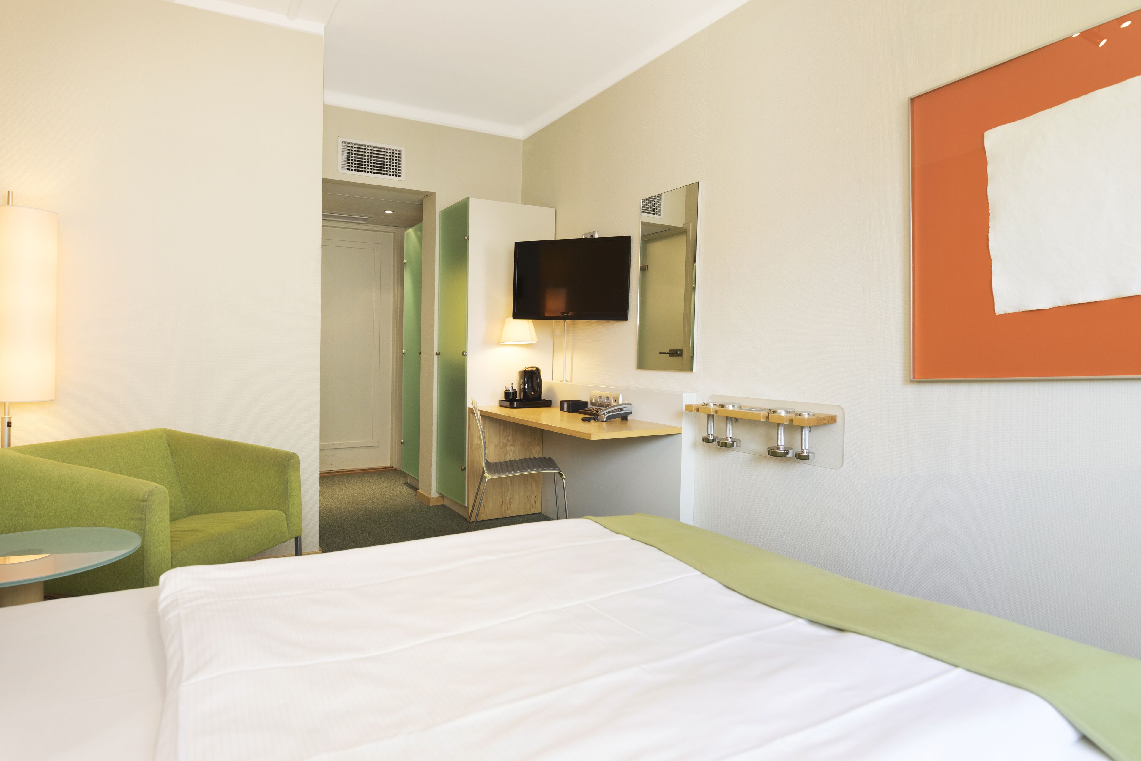 Habitación de hotel con cama, escritorio con silla, televisor montado en la pared y una mesita de noche con lámpara.