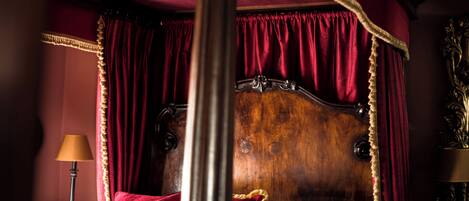 A four-poster bed with a wooden headboard, red drapery, and a nightstand with a lamp.