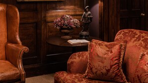 A room with a wooden panel wall, a framed portrait, a table with a lamp, a statue, and a red upholstered sofa. - The Rookery Hotel (London)