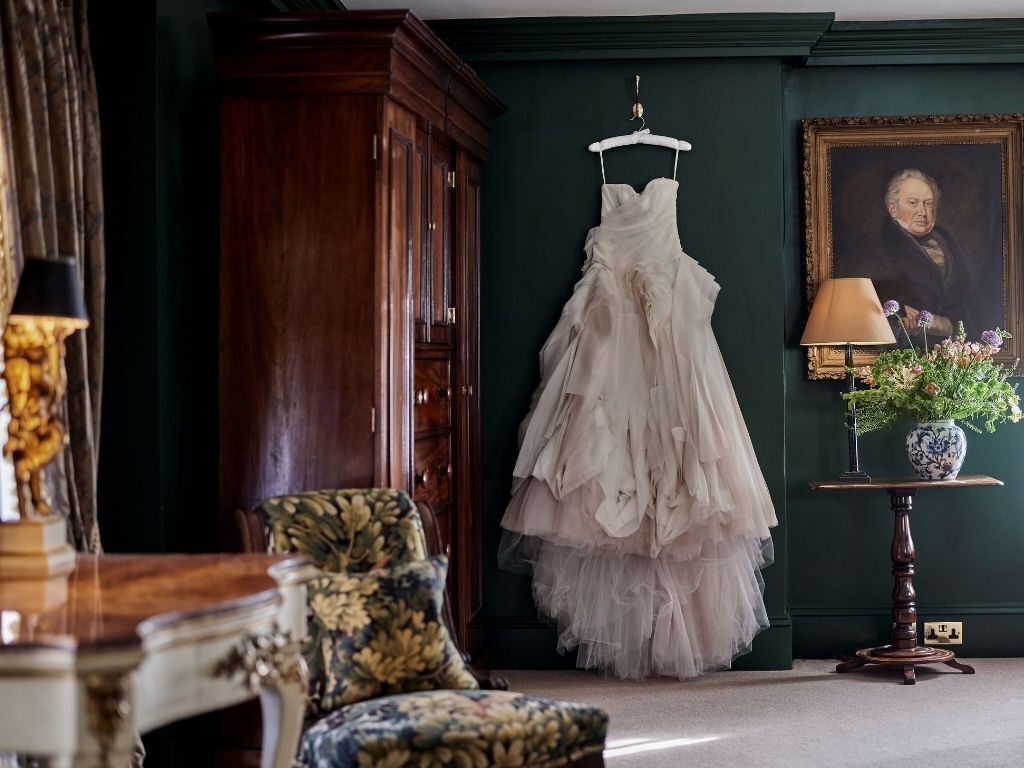 A room with a wooden wardrobe, a chair, a table with a vase, and a hanging dress.