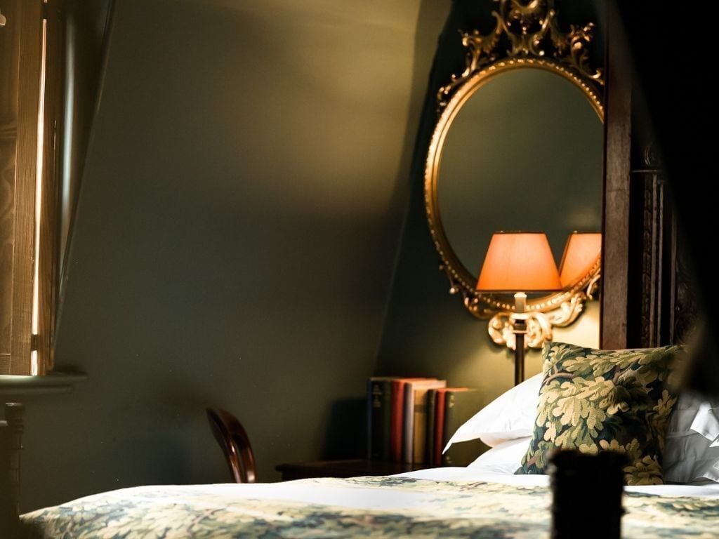 A bedroom with a bed, a bedside lamp, a mirror, and books on a nightstand.