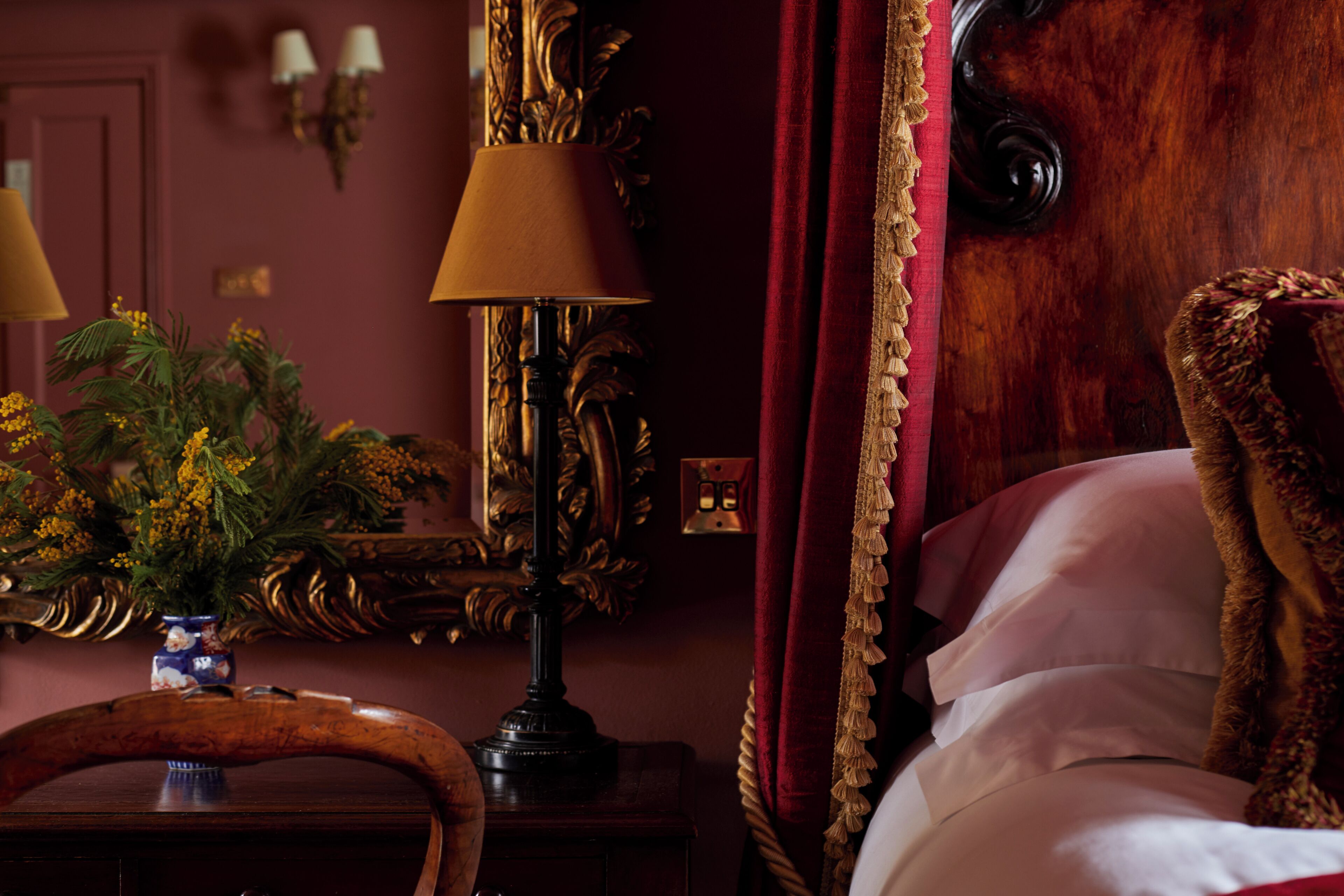 A room with a bed, a lamp, a vase with flowers, and a mirror.