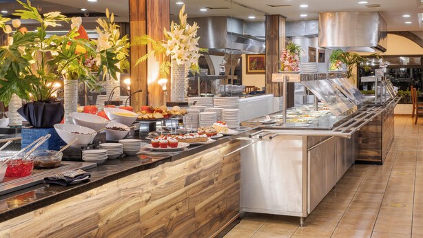 A buffet area with various food items, a stainless steel counter, and decorative plants. - Hotel Malibu (Zapopan)