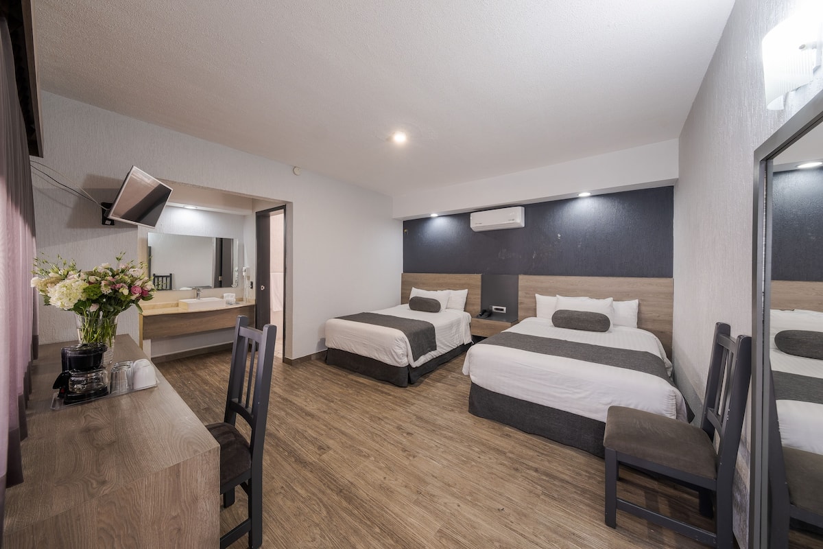 Habitación de hotel con tres camas, un escritorio con silla, un espejo y un florero con flores.