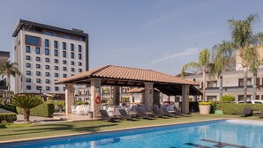 A swimming pool with lounge chairs and a gazebo, surrounded by palm trees and a multi-story building. - Hotel Malibu (Zapopan)