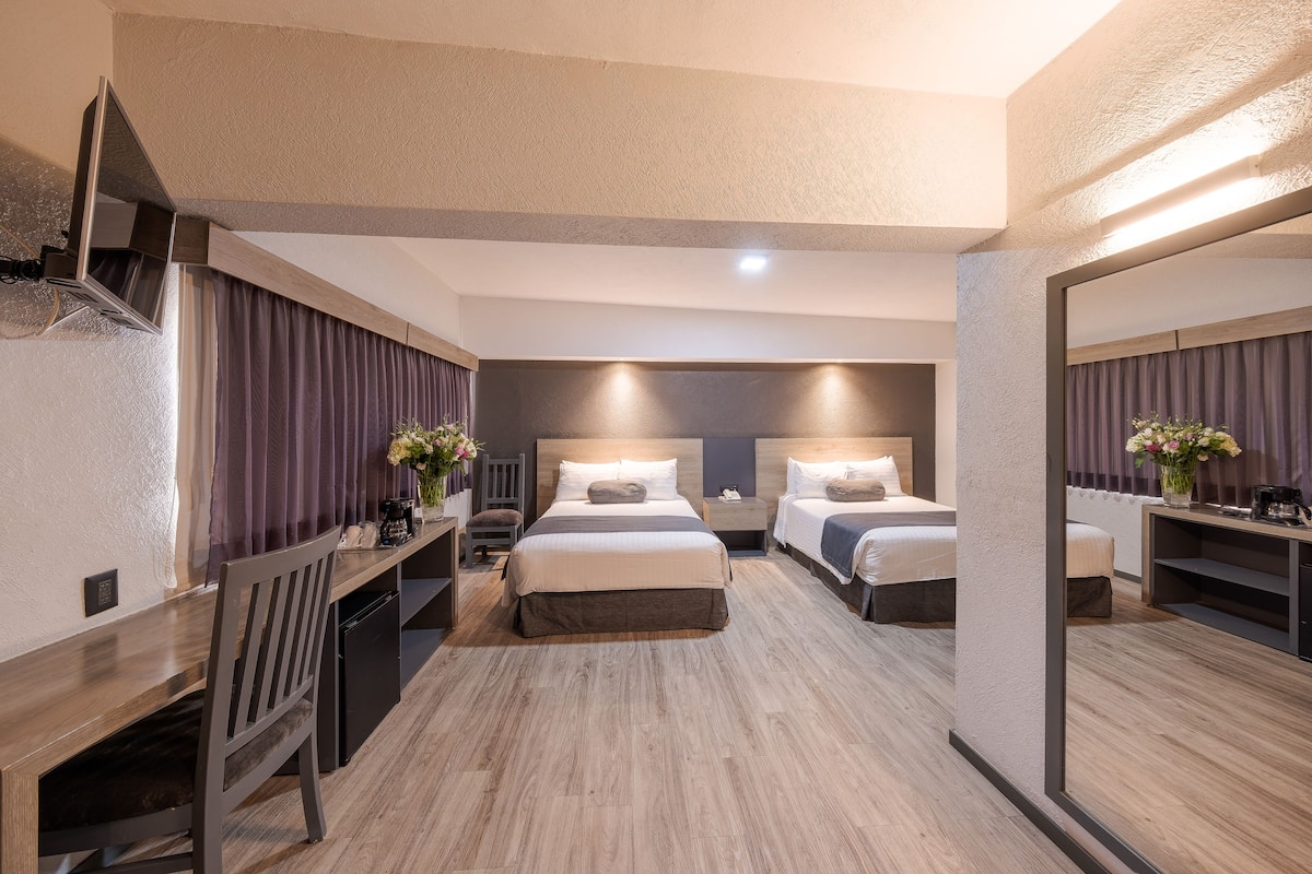 Habitación de hotel con dos camas, un escritorio y una silla. Se observa un espejo en una pared y una ventana con cortinas.