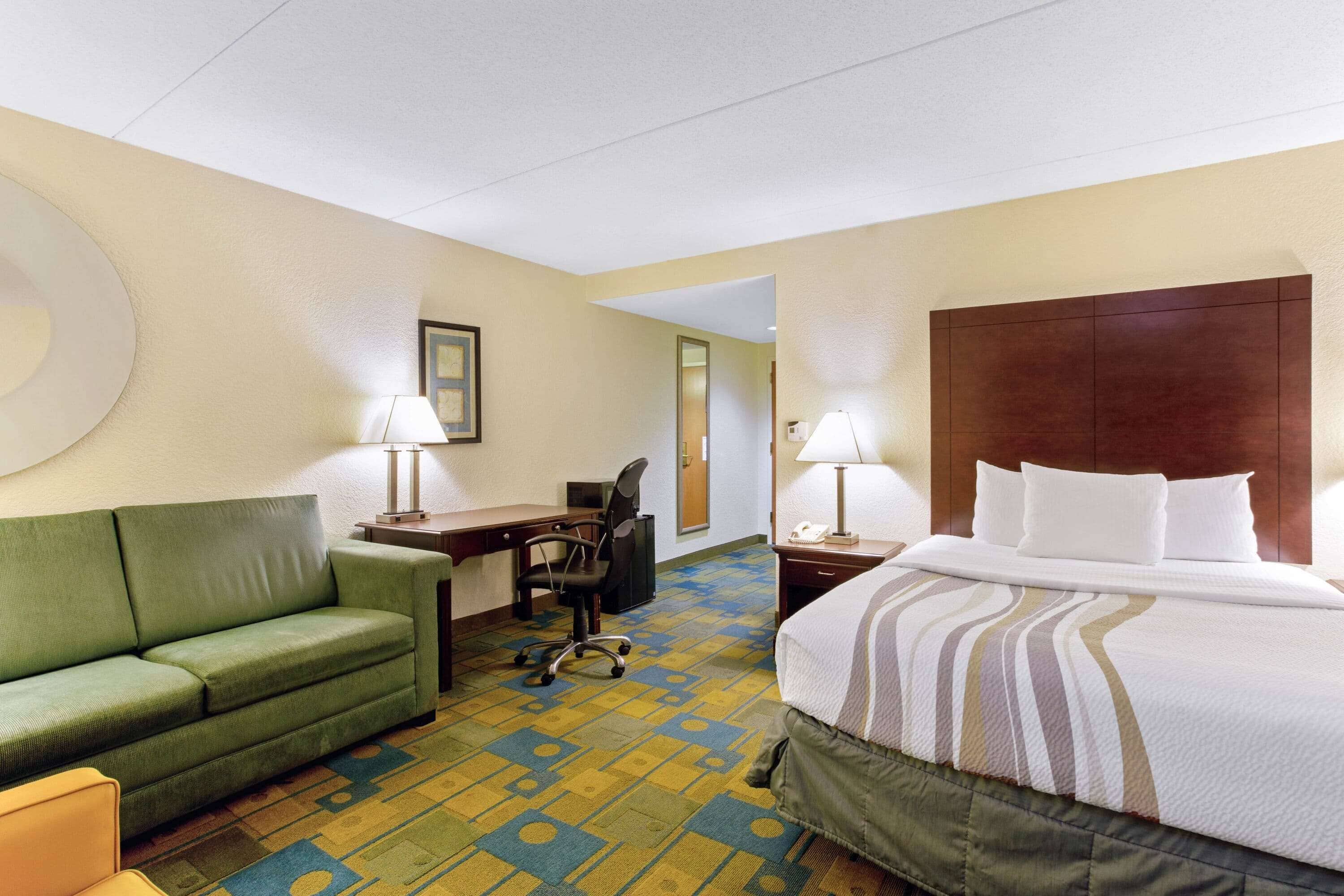 Habitación de hotel con un sofá verde, una cama con colcha a rayas, un escritorio con silla y dos lámparas.