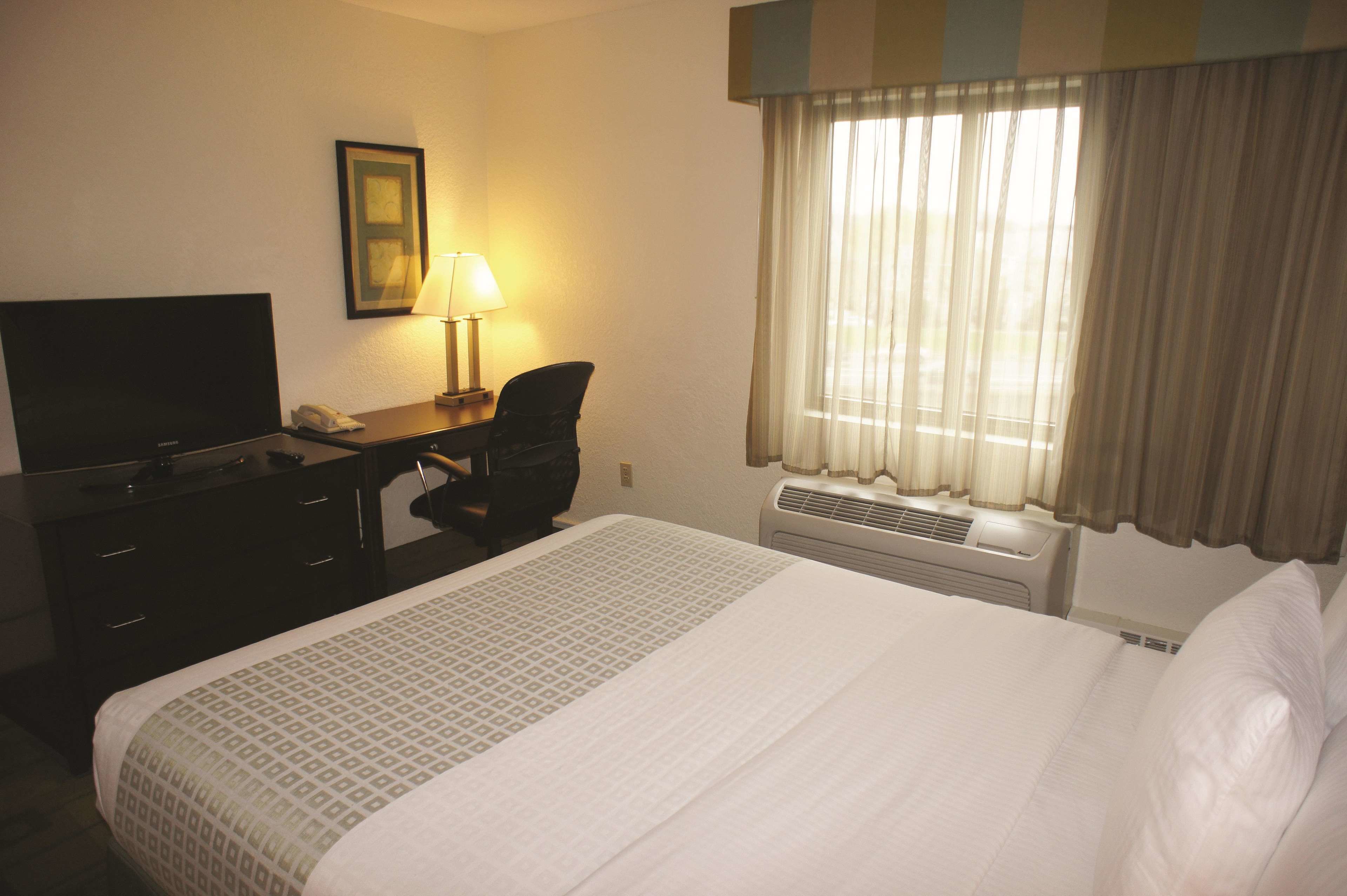 Une chambre d’hôtel avec un lit, un bureau avec une lampe, une télévision et une fenêtre avec des rideaux.