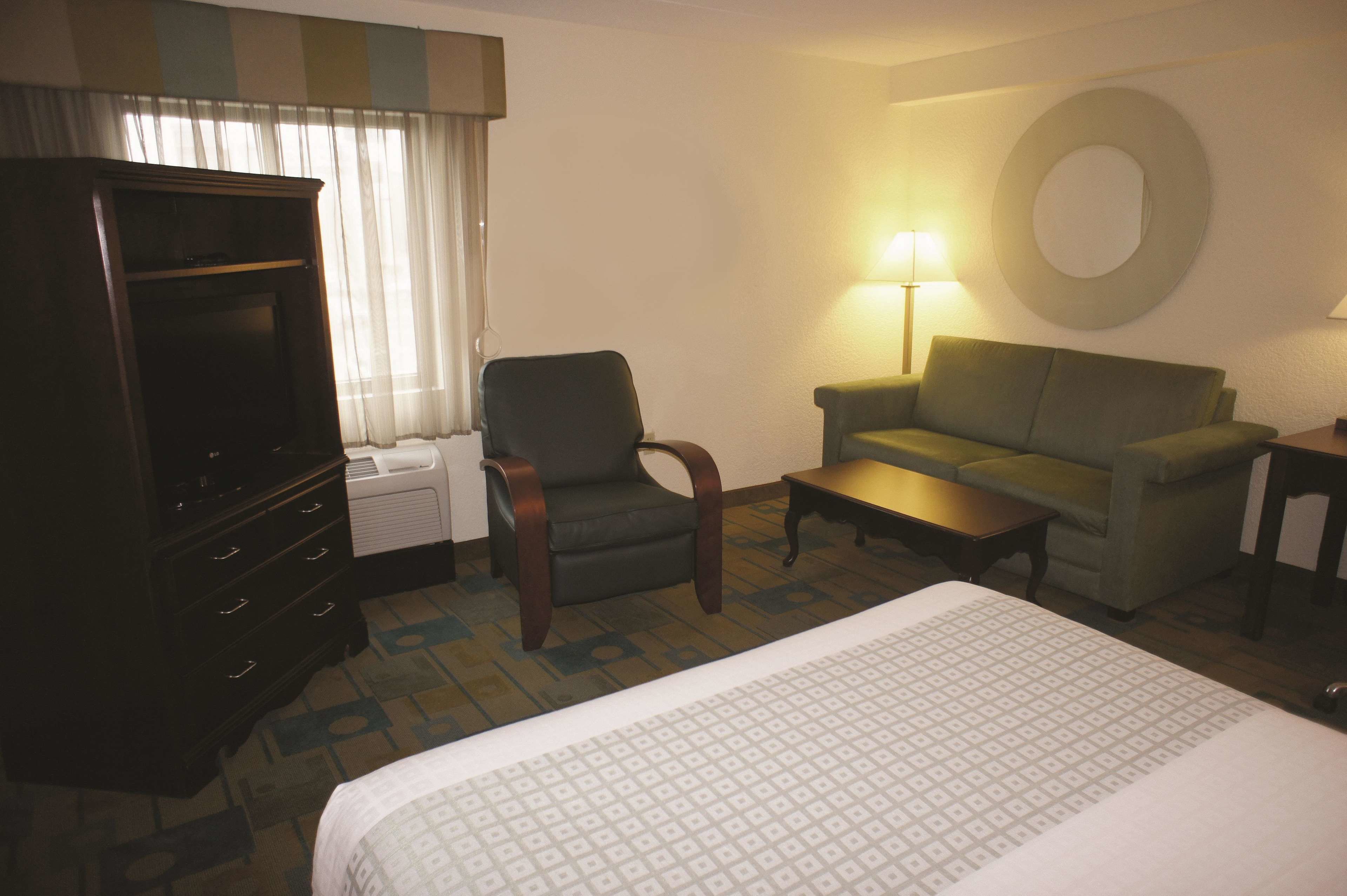 A hotel room with a bed, a TV, a sofa, a chair, a table, and a lamp.