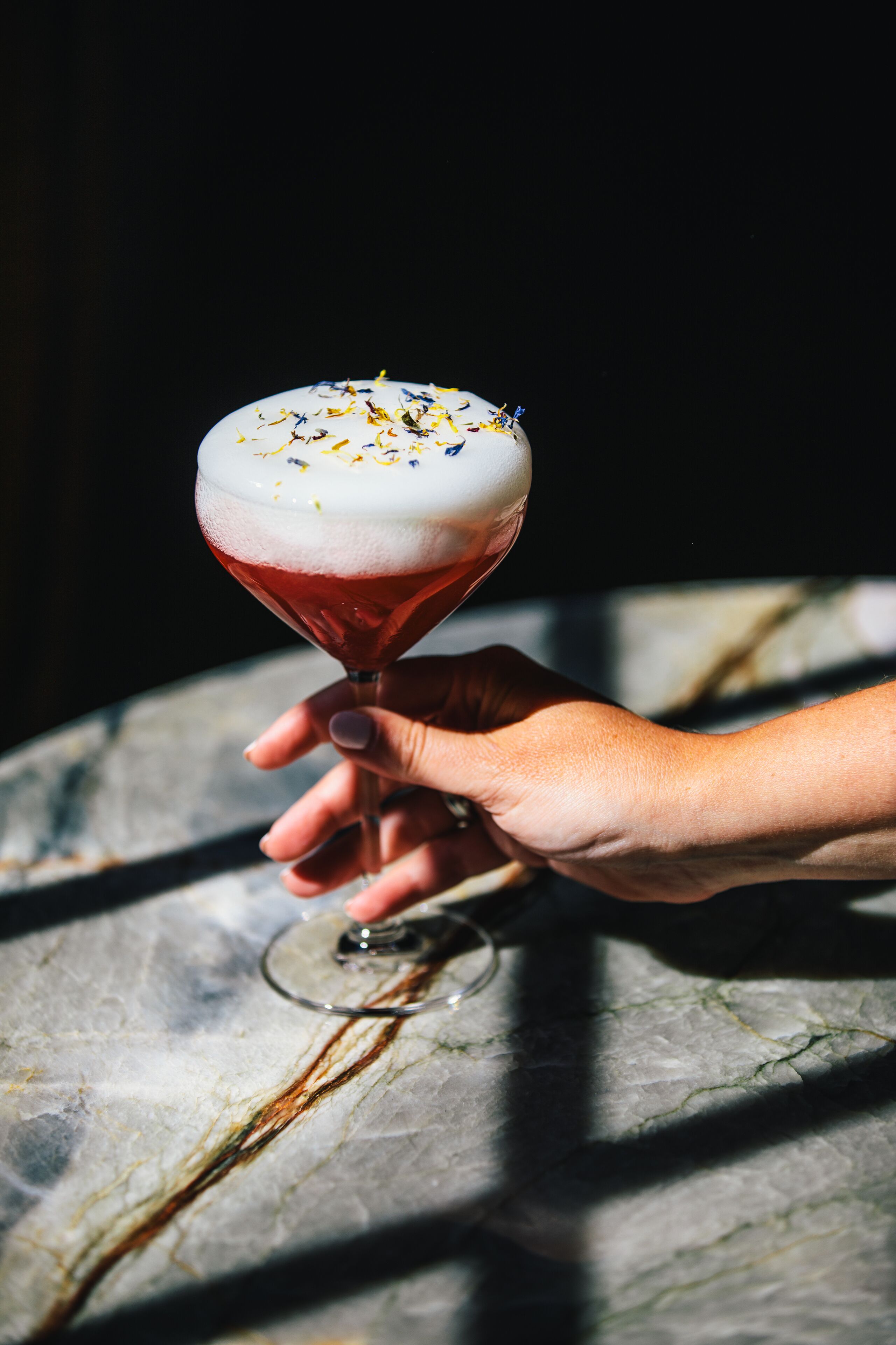 Eine Hand hält ein Cocktailglas mit schaumiger Oberfläche und farbenfroher Dekoration, vor einem Marmoruntergrund.