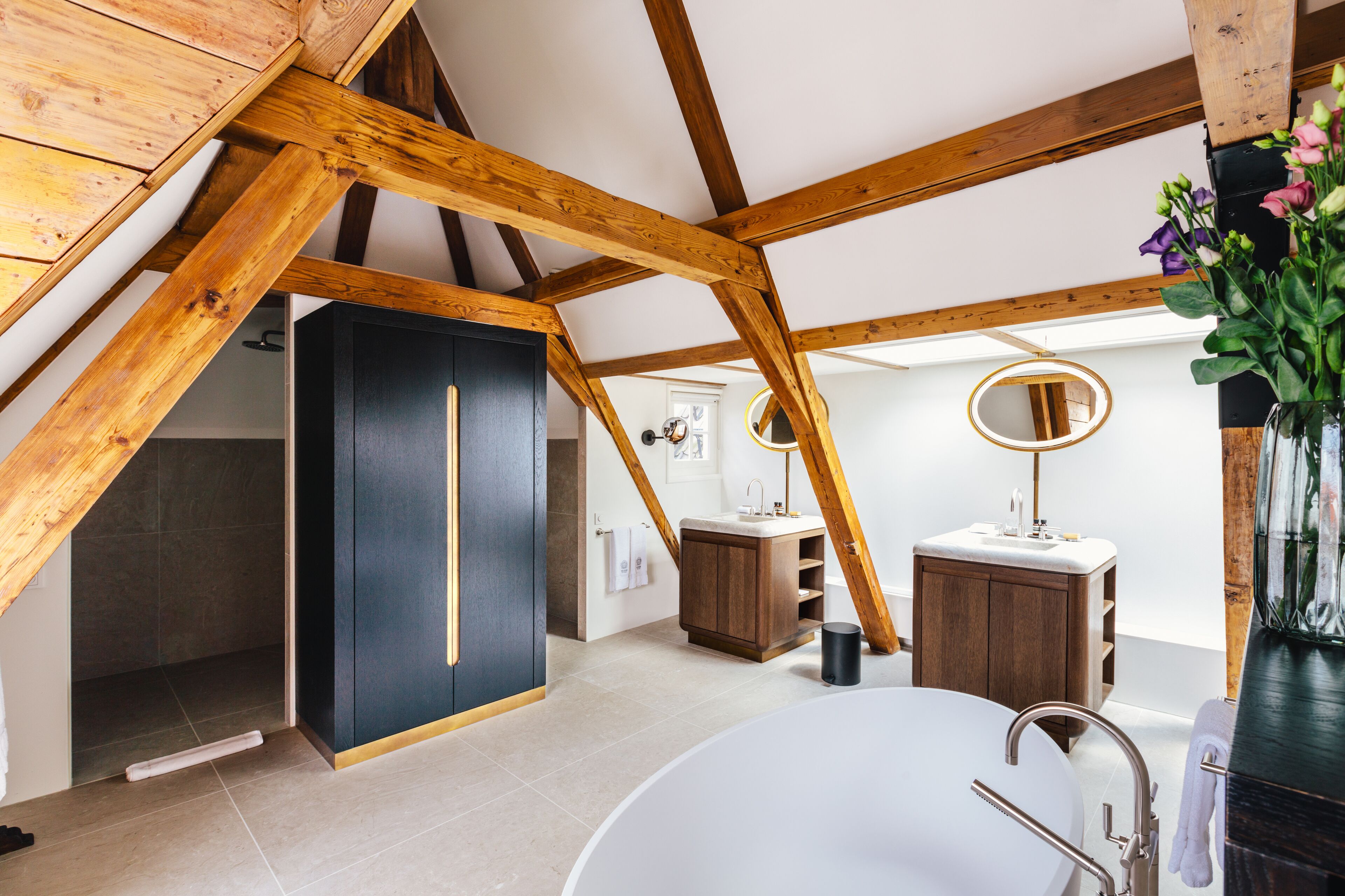 An attic bedroom with exposed wooden beams, a large window, a white bathtub, and a wooden vanity with a mirror.