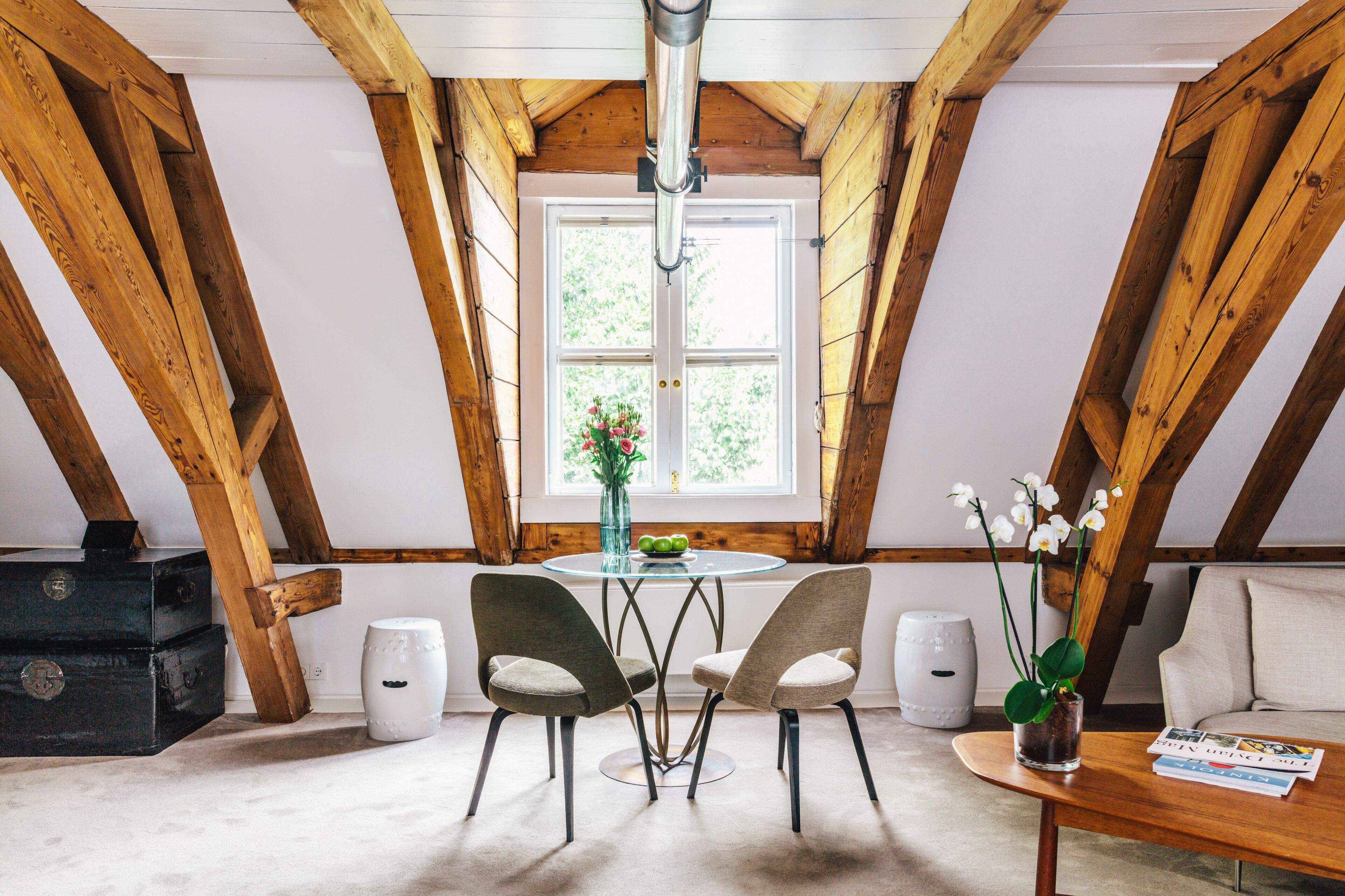 A room with a wooden ceiling, a round table with chairs, a sofa, a window with a view, and a vase with flowers.