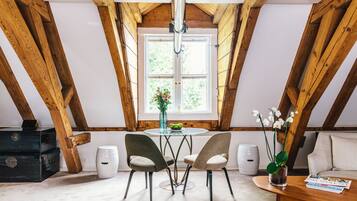 A room with a wooden ceiling, a round table with chairs, a sofa, a window with a view, and a vase with flowers.