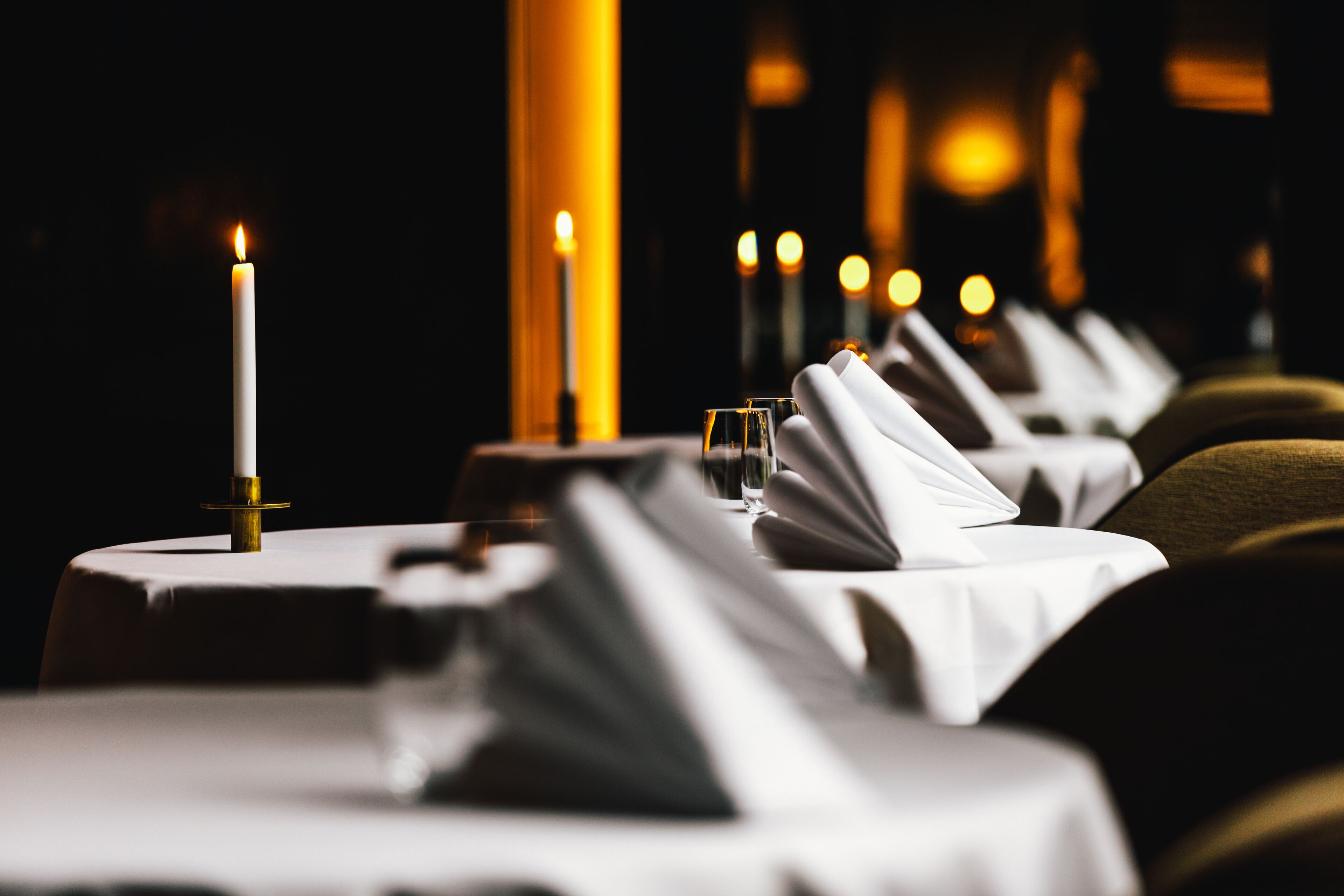 A row of tables set for dining with candles, glasses, and folded napkins.