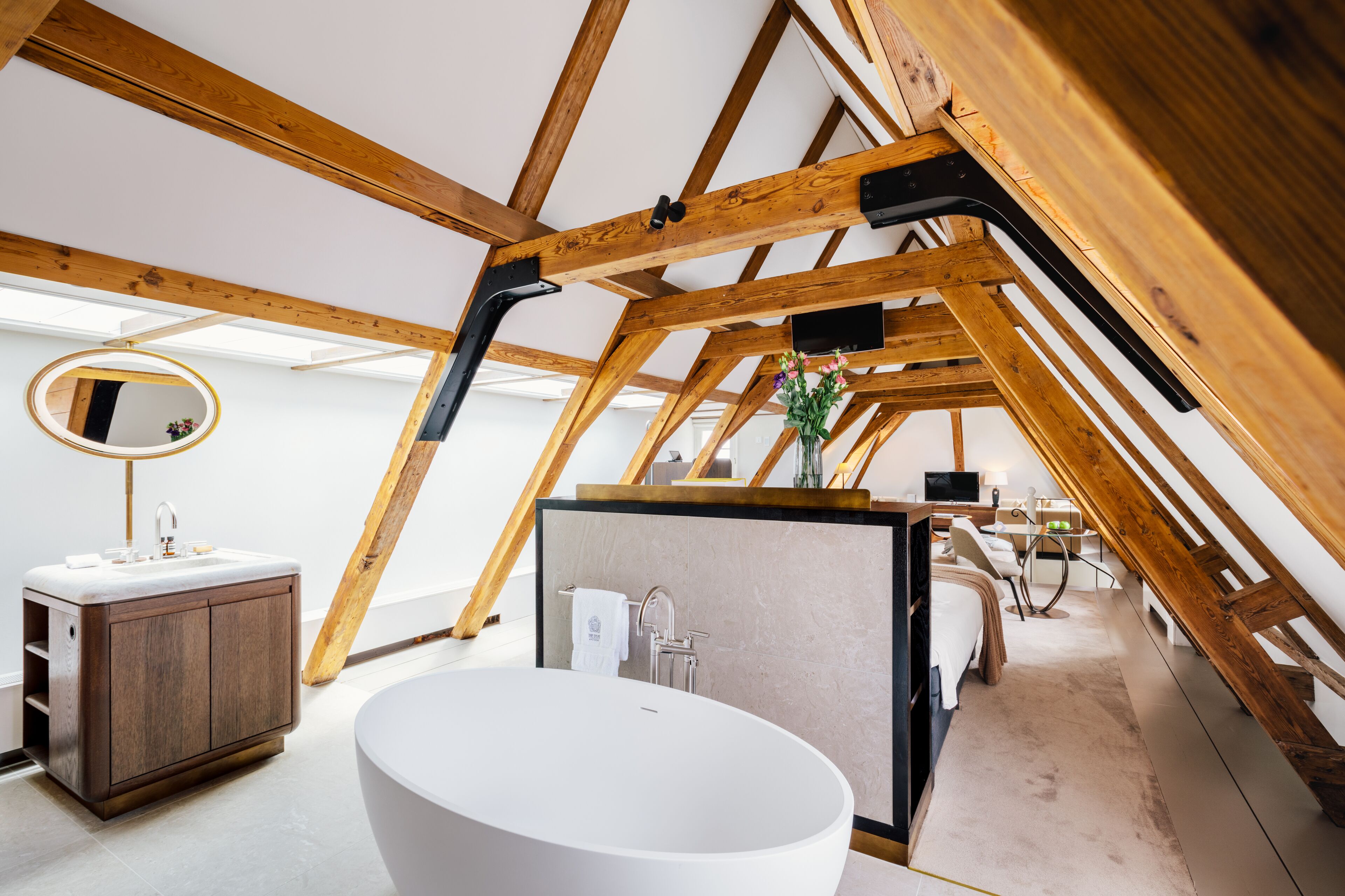 Une chambre à coucher de style loft moderne, avec des poutres en bois apparentes, une grande lucarne, un grand miroir rond, une baignoire blanche et un meuble en bois.