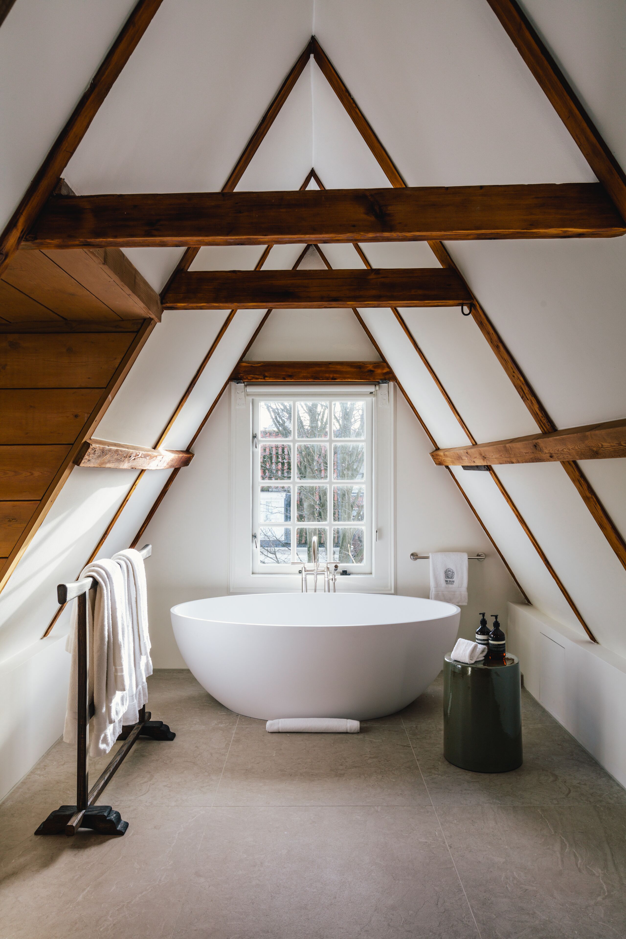 Une salle de bain aménagée dans les combles, avec une baignoire sur pieds, une fenêtre et des poutres en bois.