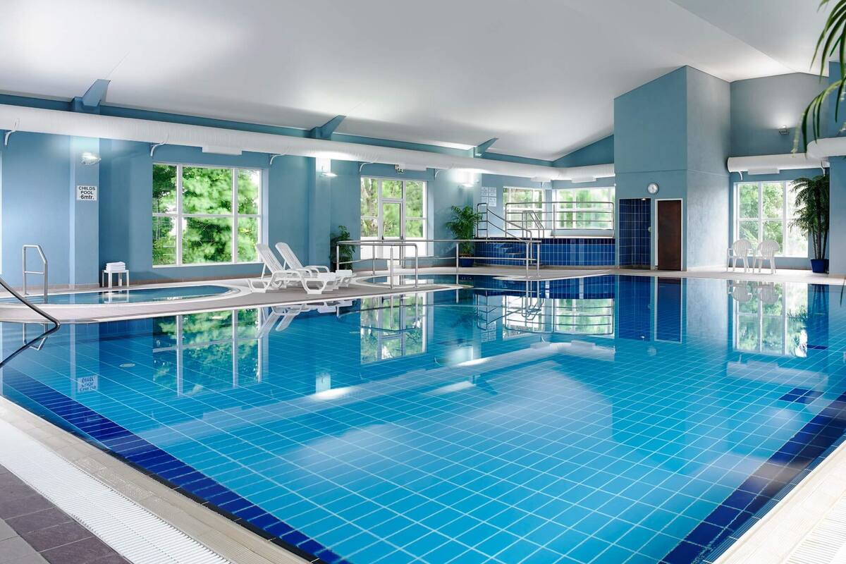 An indoor swimming pool with clear blue tiles, lounge chairs, and large windows offering views of greenery.