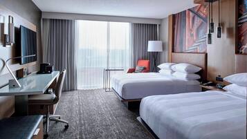A hotel room with two beds, a desk with a chair, a TV, and a large window with curtains.