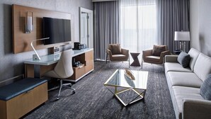 Quarto de hotel moderno com televisão de tela plana, escrivaninha com cadeira, sofá e mesa de centro de vidro.