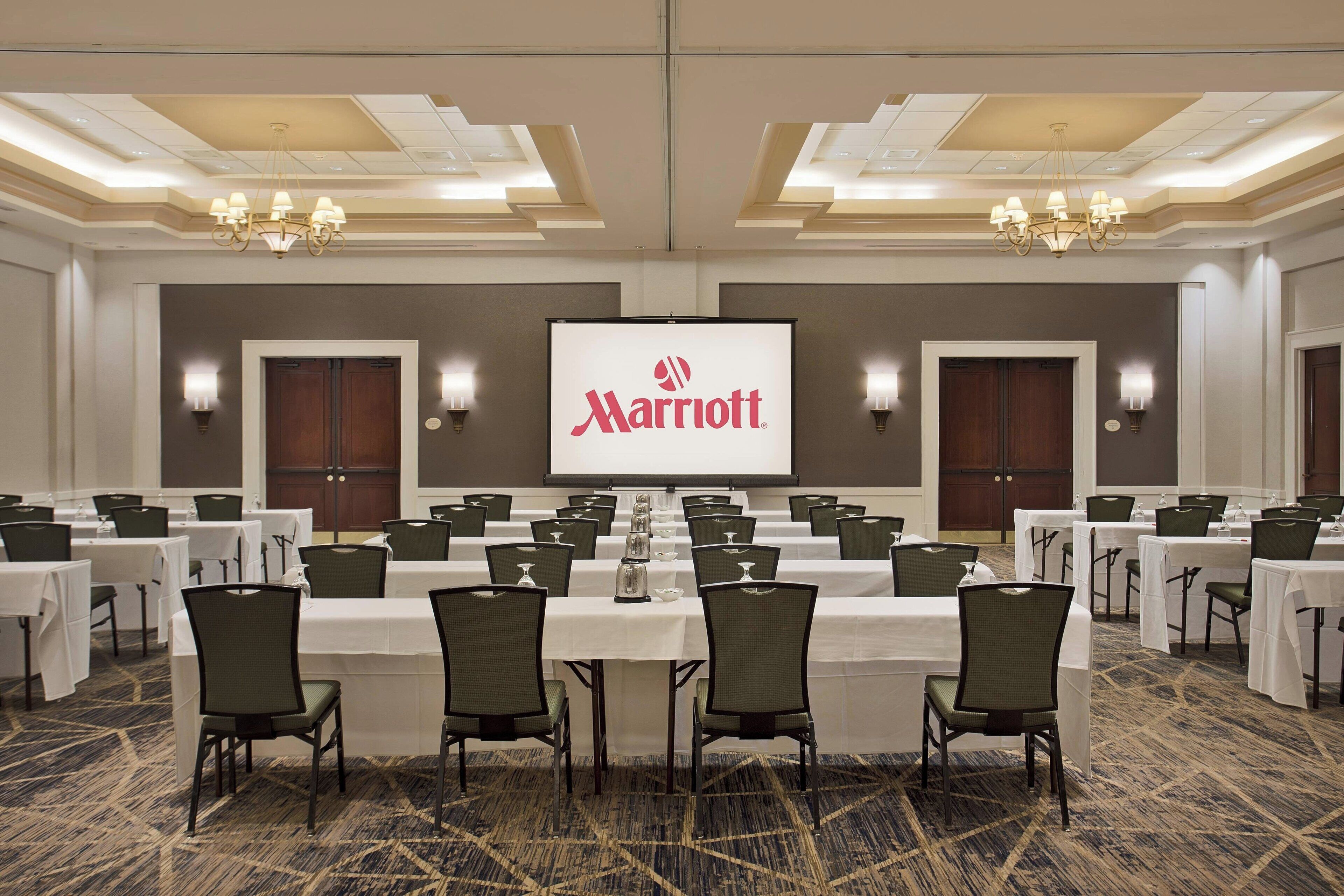 Un salón de conferencias con varias mesas y sillas, una pantalla grande que muestra el logotipo de Marriott y candelabros en el techo.