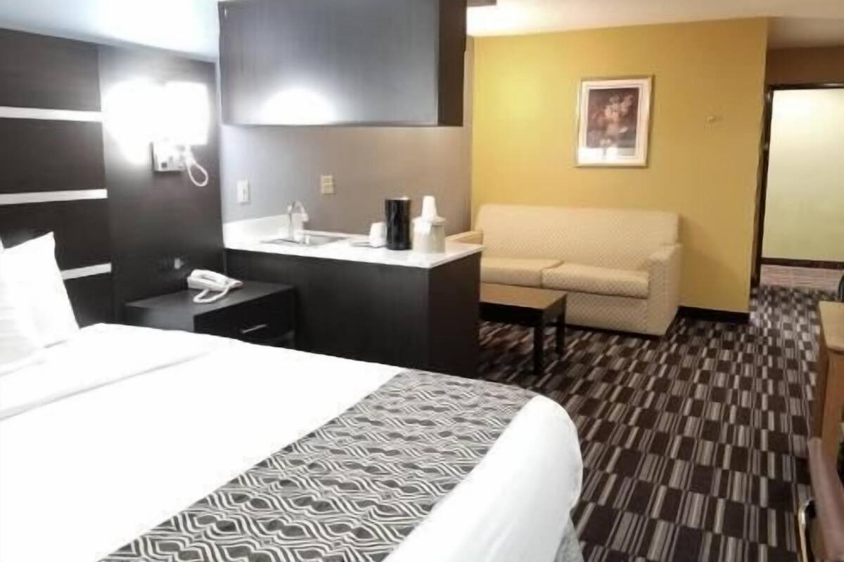 Ein Hotelzimmer mit Bett, Nachttisch mit Telefon, kleinem Schreibtisch mit Lampe, einer Couch und einem gemusterten Teppich.