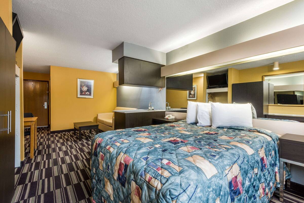 Ein Hotelzimmer mit einem großen Bett, einem Schreibtisch, einem Sessel, einem kleinen Tisch, einer Couch, einem Fernseher, einem Spiegel und einem Badezimmer mit Waschbecken und Toilette.