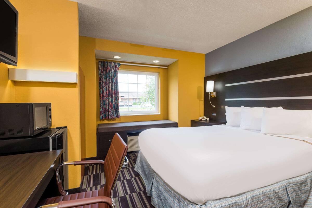 Ein Hotelzimmer mit einem großen Bett, einem Schreibtisch mit Computer, einem Fernseher und einem Fenster mit Vorhängen.