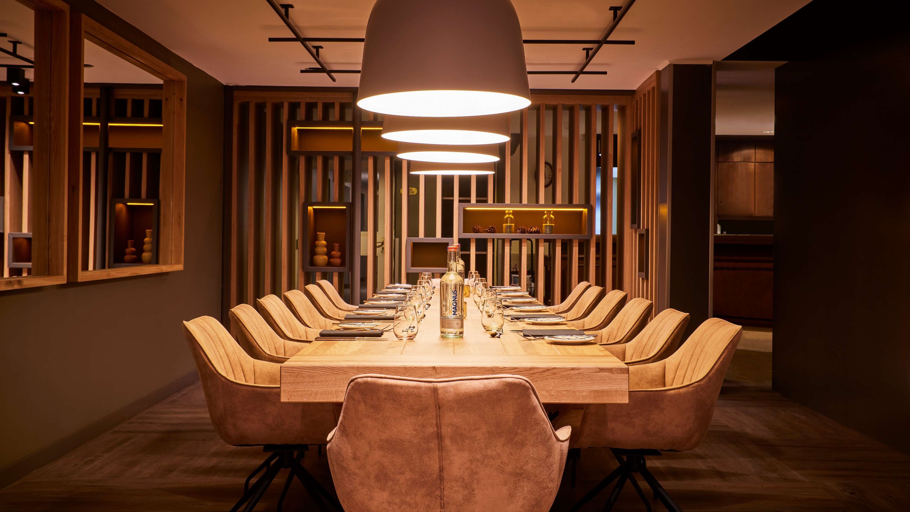 Un comedor moderno con una larga mesa de madera, varias sillas y una botella de MAGNUS en el centro.