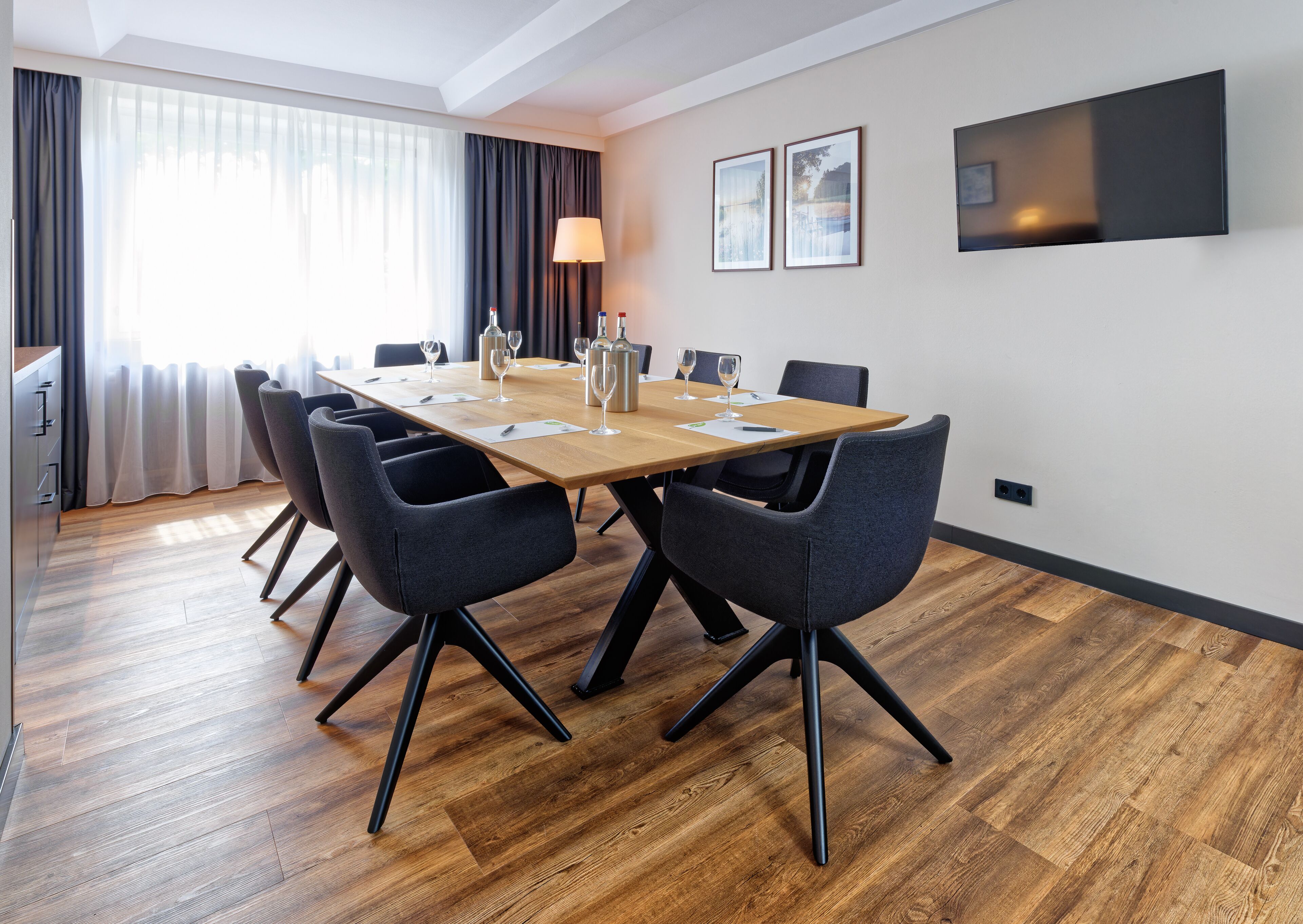 A conference room with a wooden table, six chairs, a lamp, and two framed pictures on the wall.