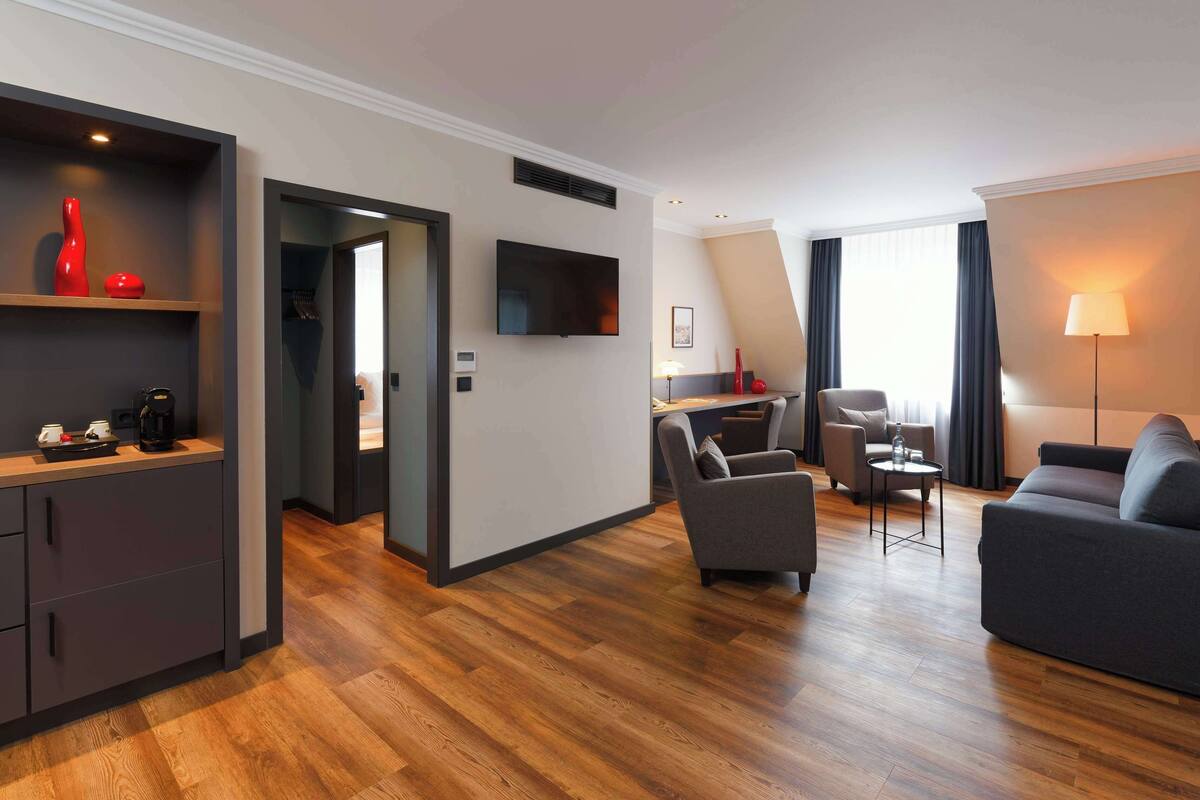 Una habitación de hotel moderna con piso de madera, un sofá gris, una pequeña mesa redonda, un sillón gris, un televisor montado en la pared y una cafetera sobre la encimera.