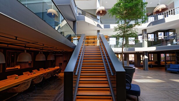 Ein modernes Interieur mit einer zentralen Treppe, Holztischen und Sitzbereichen.