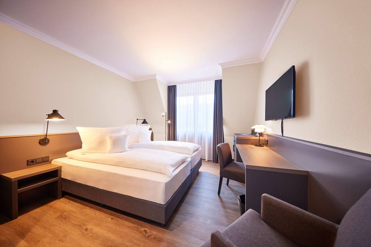 Habitación de hotel con cama, mesita de noche, lámpara, silla, escritorio y televisor montado en la pared.