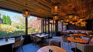 Ein Restaurant mit Holzbalkendecke und großen Fenstern, das einen Blick auf einen Garten bietet. Tische und Stühle sind für das Essen bereitgestellt.