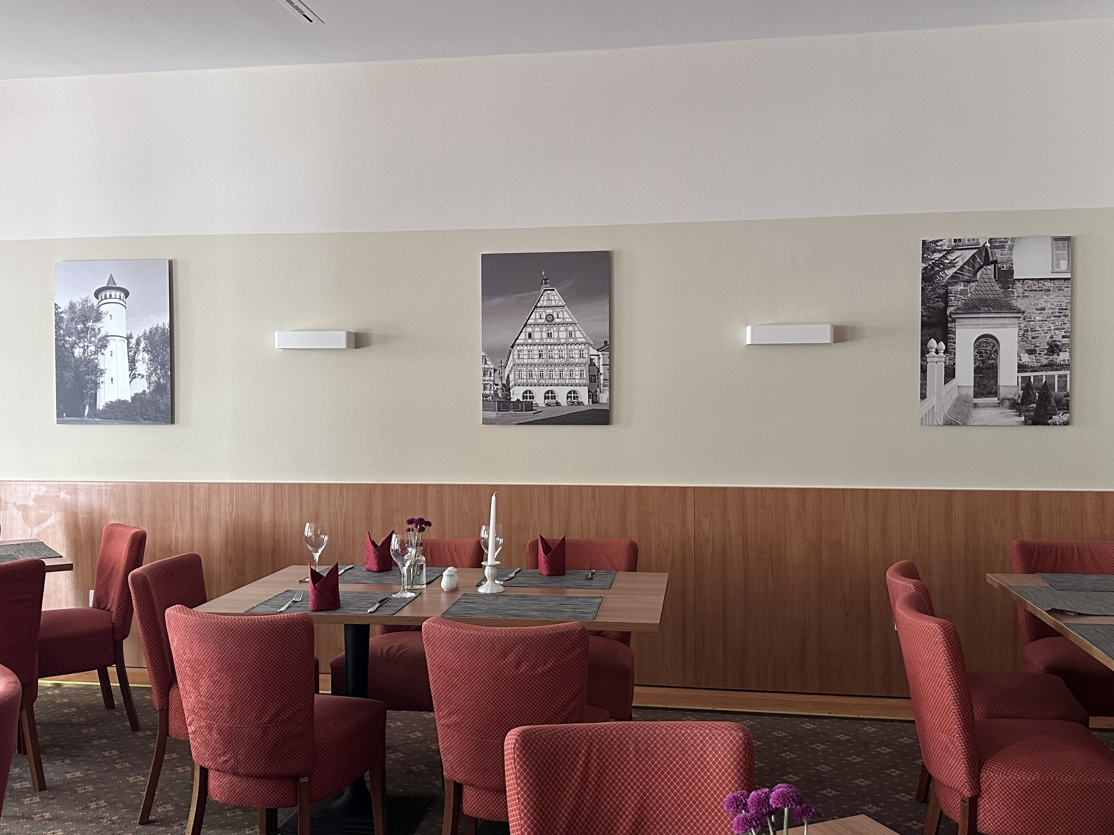 Un coin repas avec des chaises à revêtement rouge, une table en bois mise avec des couverts, et trois photographies en noir et blanc accrochées au mur.
