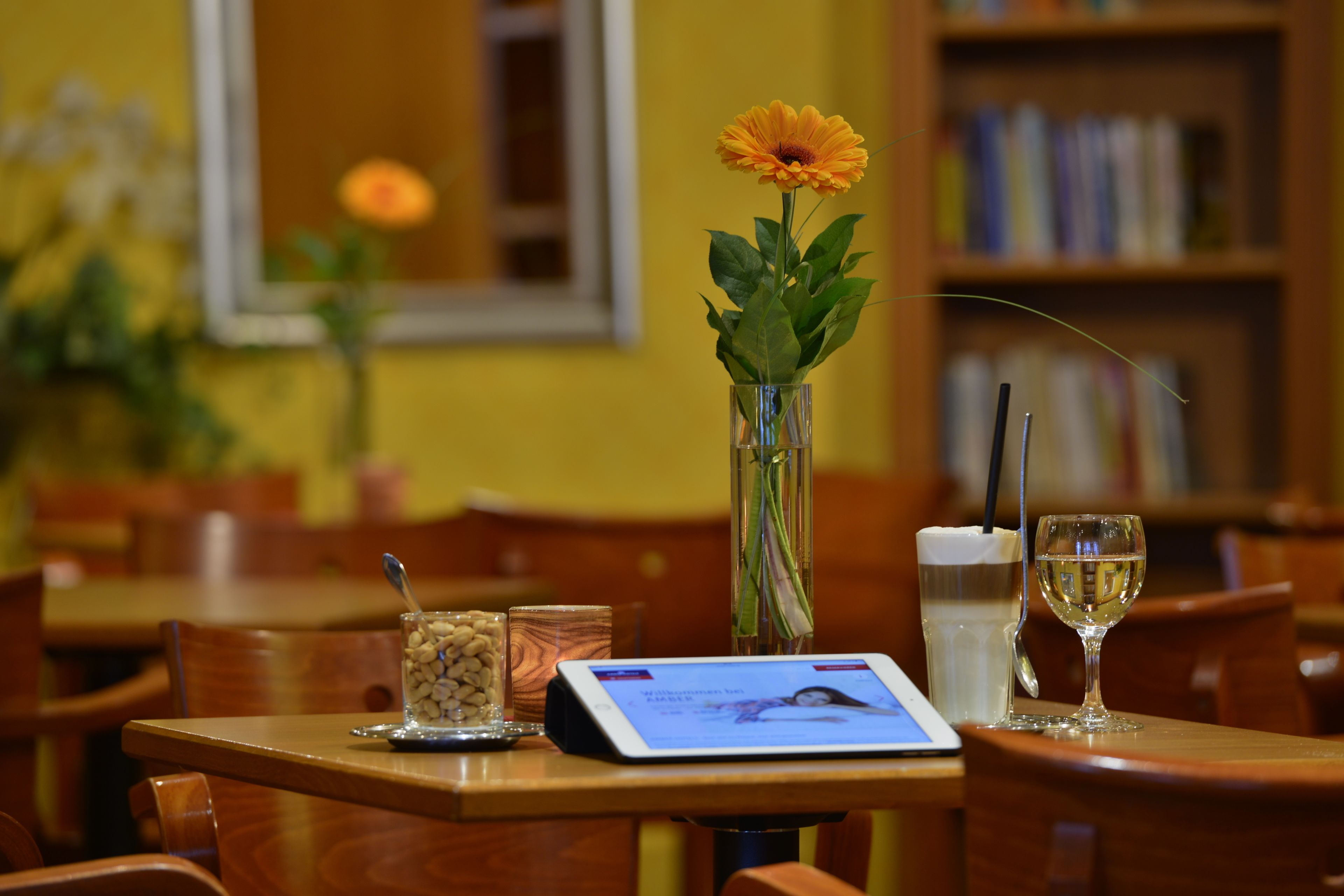 Una mesa de comedor con una tableta, una copa de vino blanco, un florero con una flor y una taza de café.