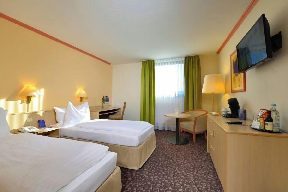 A hotel room with two beds, a desk, a chair, a television, and a small table with a lamp.