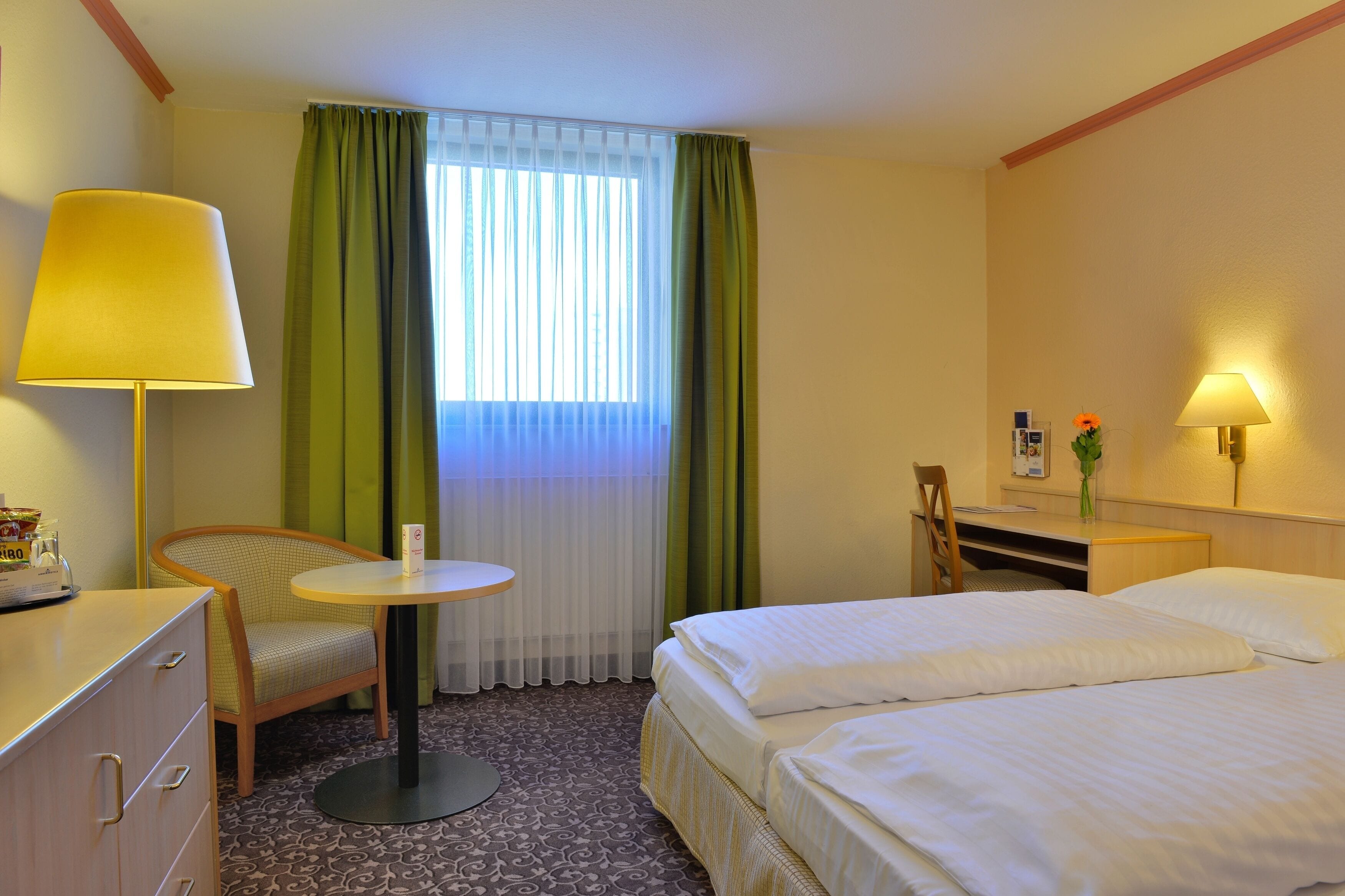 A hotel room with two beds, a desk, a chair, a lamp, and a window with curtains.