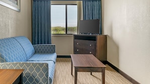 A hotel room with a sofa, a wooden table, a TV on a stand, and a window with curtains. - Best Western Port Huron Blue Water Bridge (Port Huron)