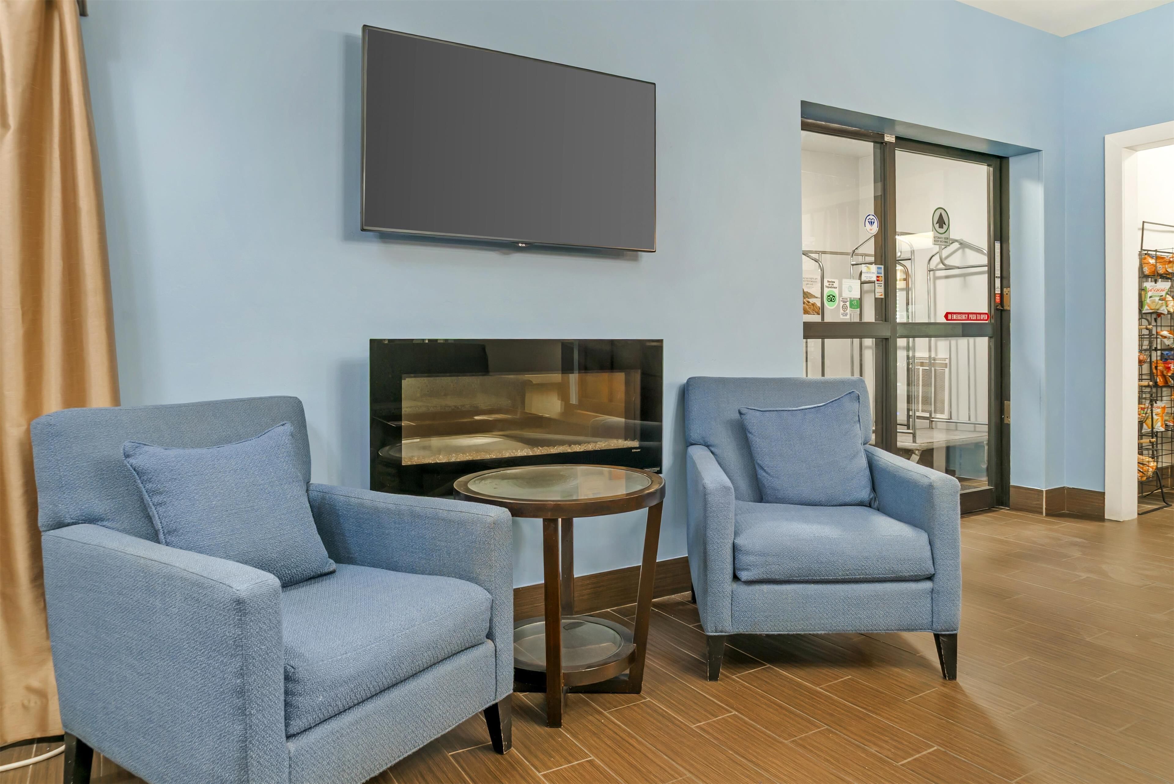 A room with two blue armchairs, a small round table, a flat-screen TV mounted on the wall, and a glass door leading to a balcony with a railing and a sign.