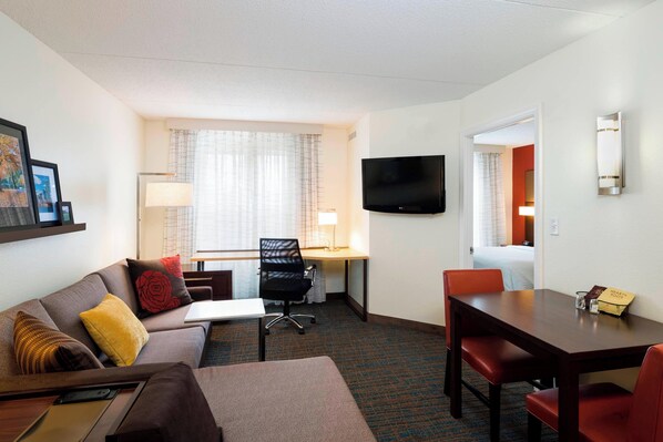 Habitación de hotel con sofá, escritorio, mesa de comedor con sillas, televisor montado en la pared y ventana con cortinas.