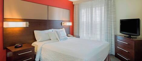 Una habitación de hotel con una cama grande, una mesita de noche con lámpara, un televisor en un aparador y una ventana con cortinas.