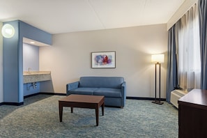 A hotel room with a blue sofa, a small table, a painting on the wall, a lamp, and a window with curtains.