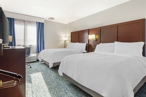 Quarto de hotel com duas camas, uma escrivaninha, uma cadeira, um abajur e uma janela com cortinas.