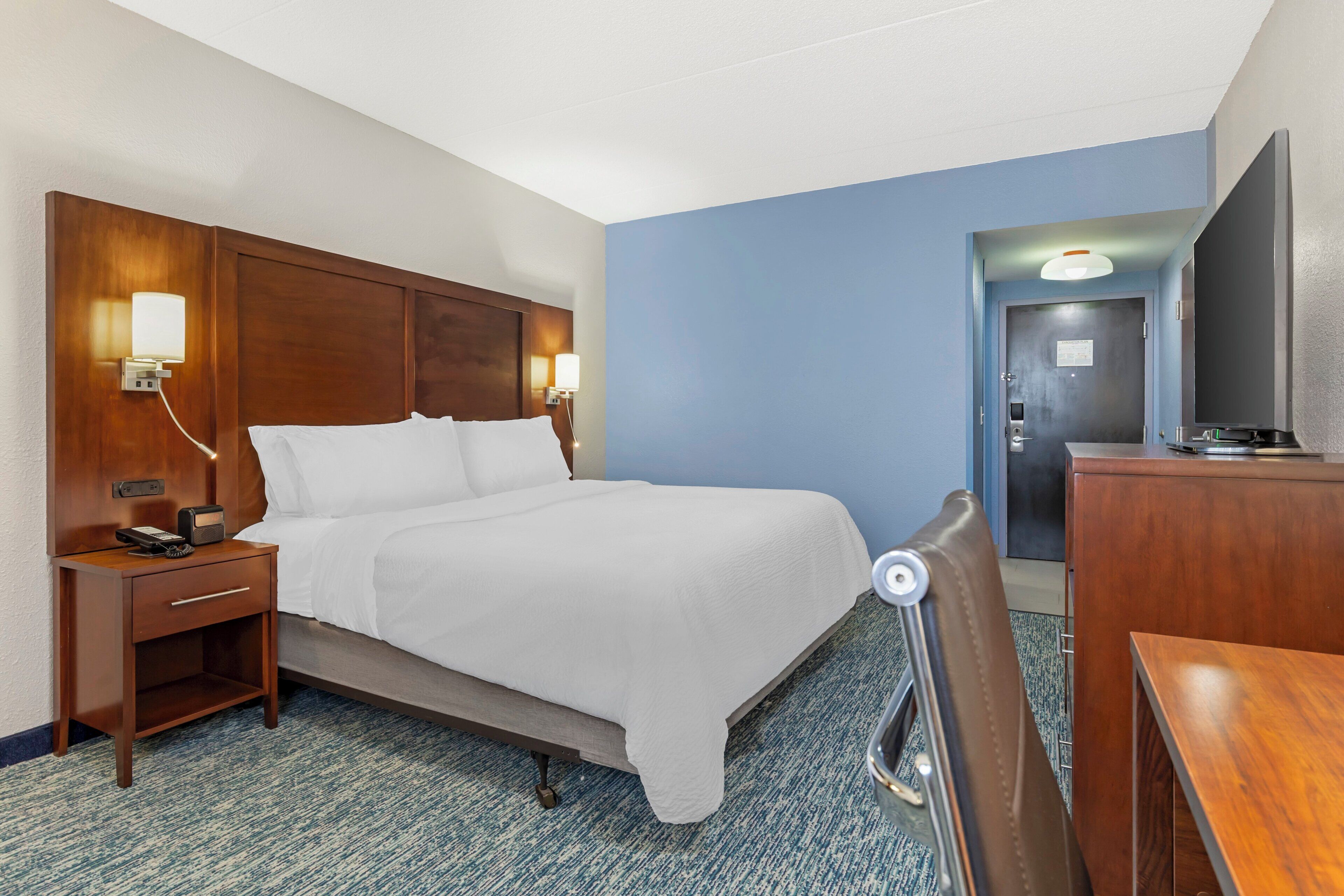 A hotel room with a bed, bedside table, lamp, and a desk with a chair.