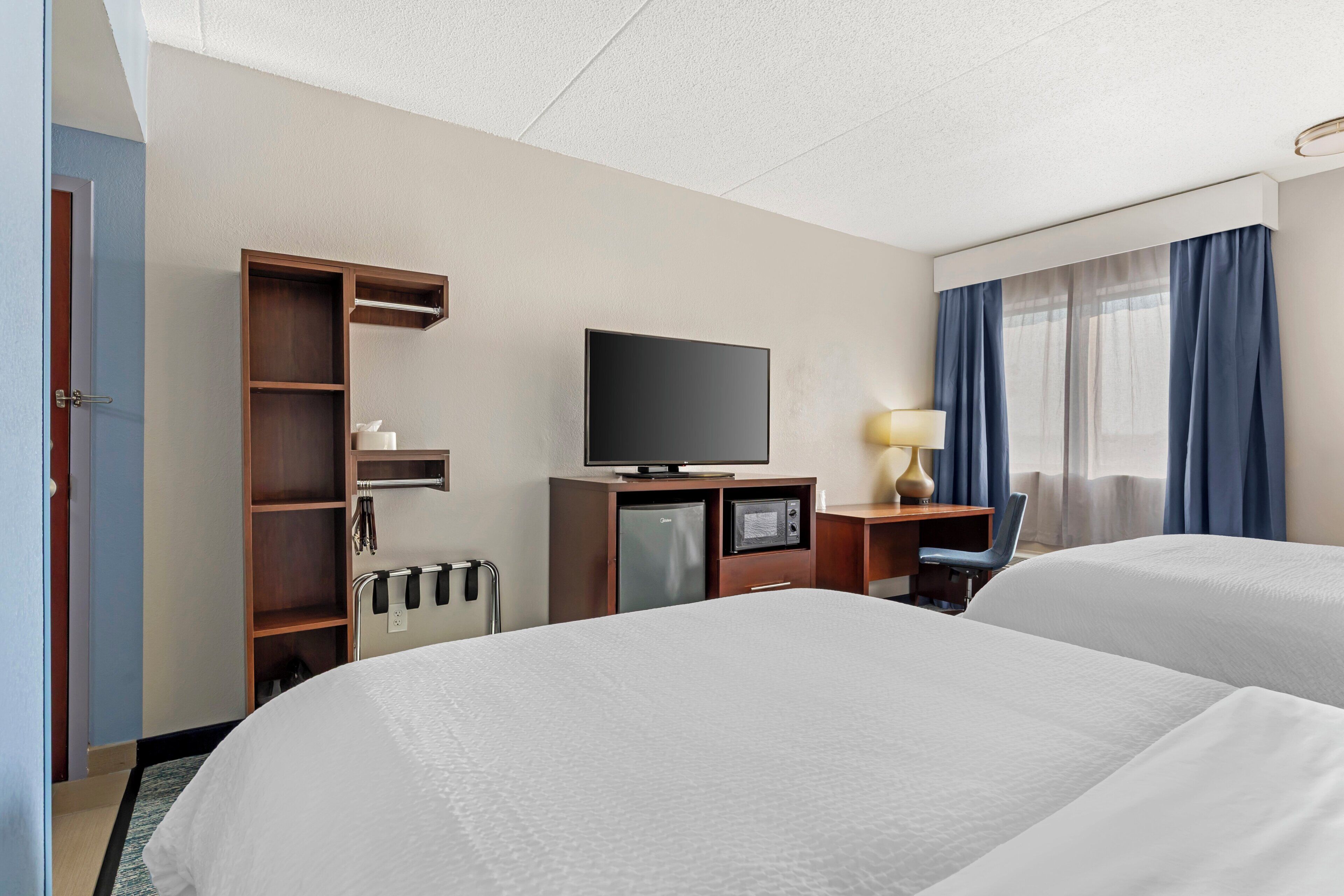 Ett hotellrum med två sängar, en tv, ett skrivbord med en lampa, en minibar och ett fönster med gardiner.