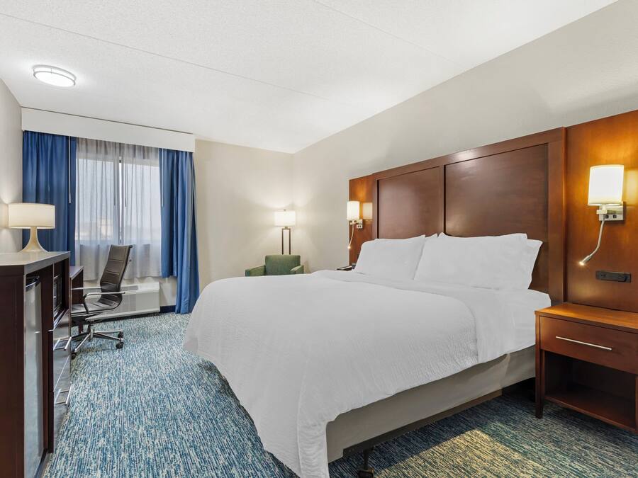 A hotel room with a bed, a desk, a chair, a nightstand, and a lamp.